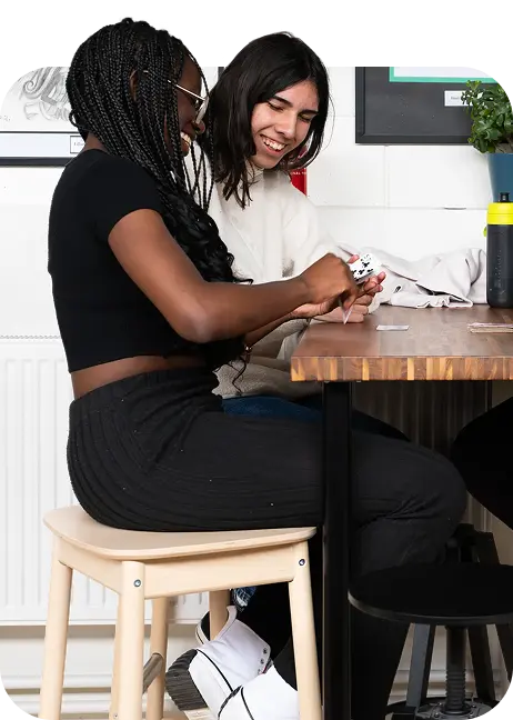 Two women sit next to each other at a wooden table in the ELC, smiling and playing cards. One woman has plaited hair and wears glasses, while the other has dark hair and wears a light jumper.