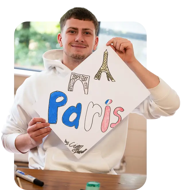 A young man in a white hoodie smiles and holds up a sign that says Paris with colourful letters, ELC, and hand-drawn sketches of the Eiffel Tower and Arc de Triomphe. A green marker and pen are on the table in front of him.