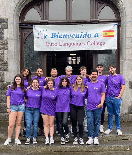 A group of thirteen young adults in matching purple ELC T-shirts stand smiling on stone steps in front of a building, beneath a banner that reads “Bienvenido a Euro Languages College,” with a Spanish flag on the right.