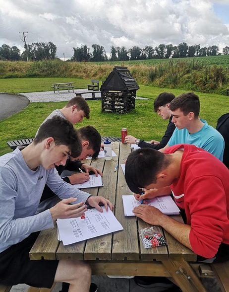 Six young men sit at a wooden picnic table outdoors, focused on ELC papers in front of them. The setting is grassy with trees and a cloudy sky in the background.